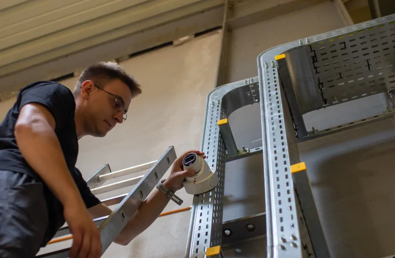Instalator ustawia kamerę CCTV przy metalowej trasie kablowej w hali.