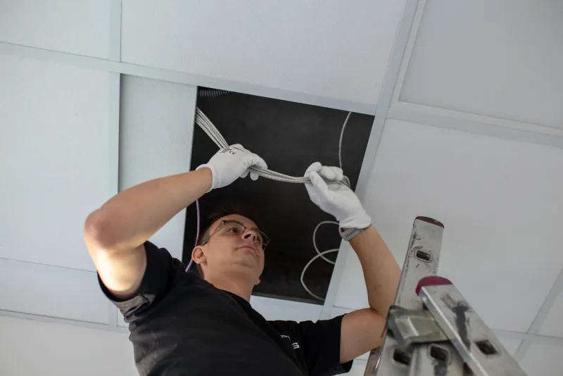 Instalator w rękawicach układa wiązkę przewodów nad sufitem podwieszanym.